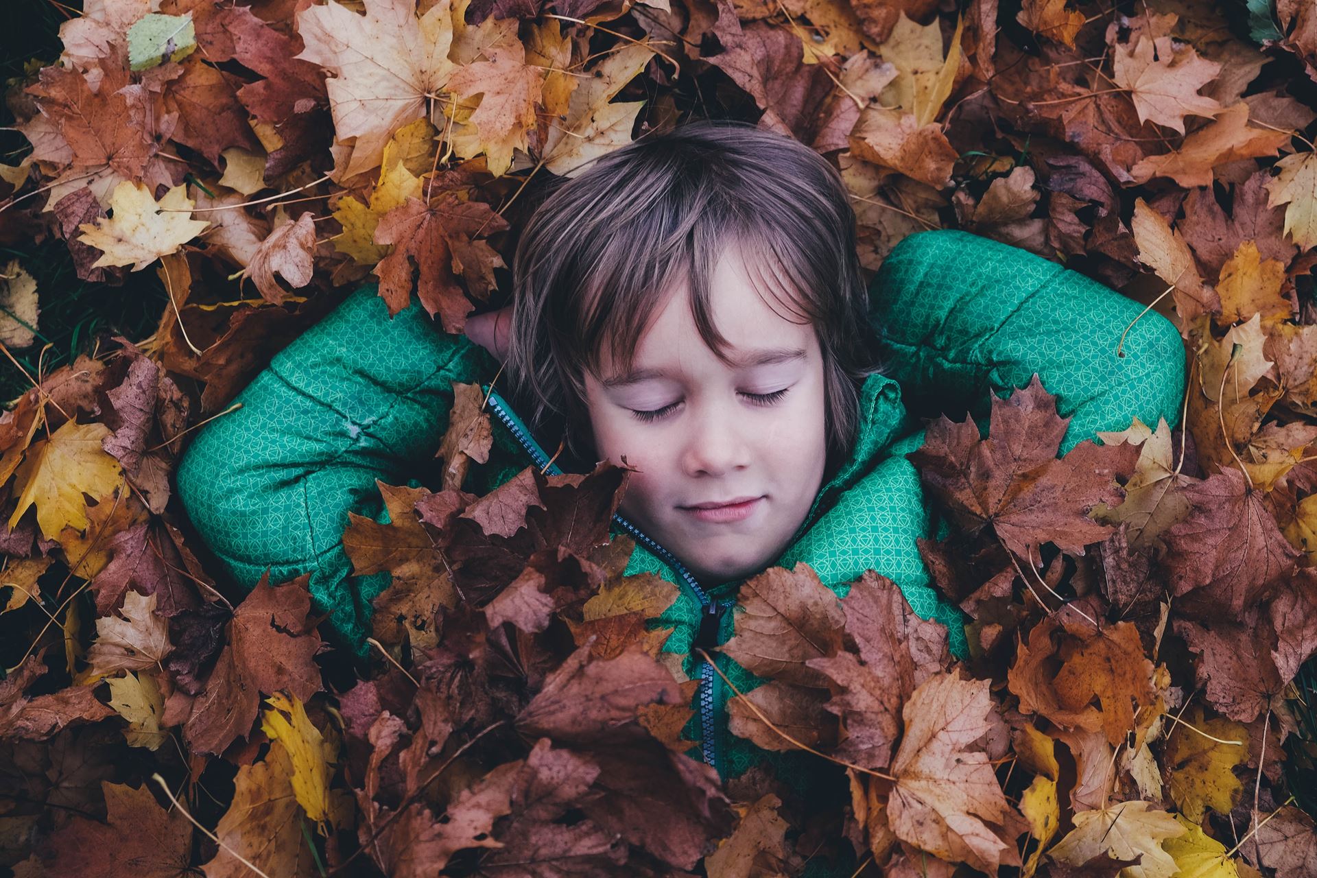 A child in a green coat laying in brown leaves with their eyes closed